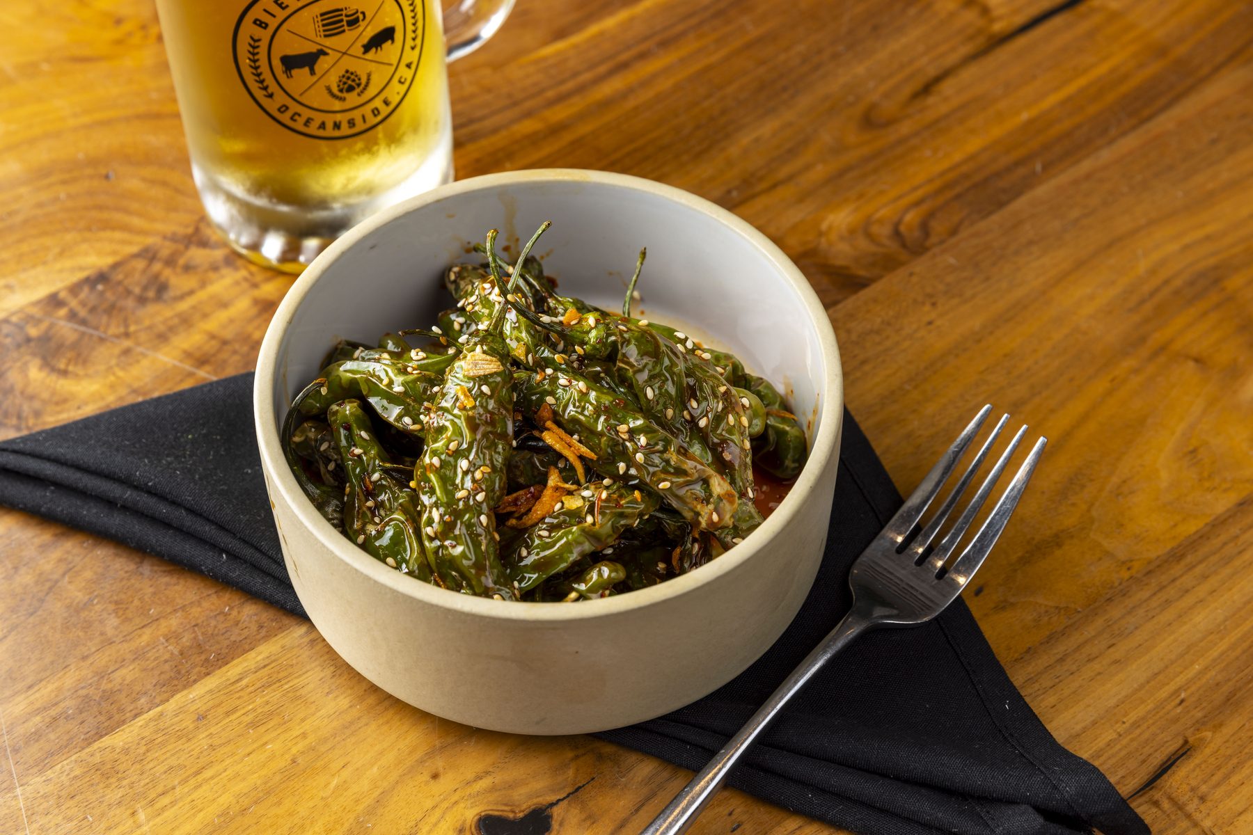 Blistered shishito peppers in a bowl beside a Bier Garden lager